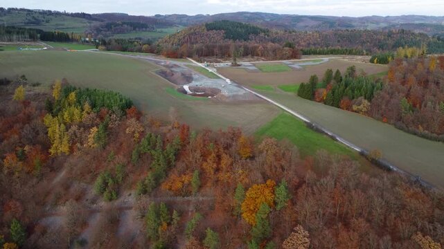 Drohnenflug mit Aufw&auml;rtsbewegung &uuml;ber Windkraft-Baustelle in herbstlicher Mittelgebirgslandschaft &ndash; Blick auf Windr&auml;der im Bau, Baustellenfahrzeuge und Schotterstra&szlig;e, Symbol f&uuml;r Energiewende