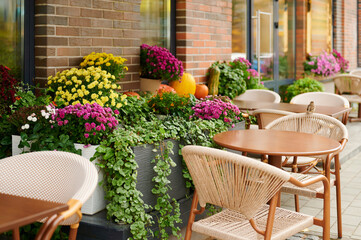 Fototapeta premium outdoor cafe scene with empty wicker chairs and wooden tables surrounded by vibrant flowers and foliage. charming seating arrangement evokes cozy, relaxed ambiance in urban setting