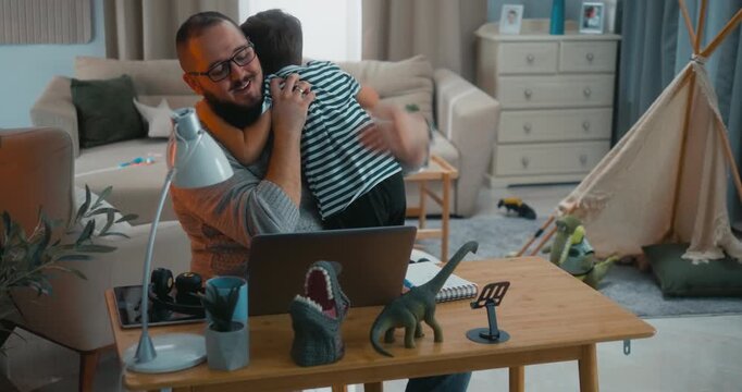 Busy father working remotely on his laptop gets hugged and lifted by his son, who invites him to play. Dad closes the computer and joins him, showing family first and positive work-life balance.