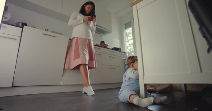 Mother standing in kitchen looking at phone while baby sits on floor exploring surroundings, modern parenting moment showing multitasking and early development