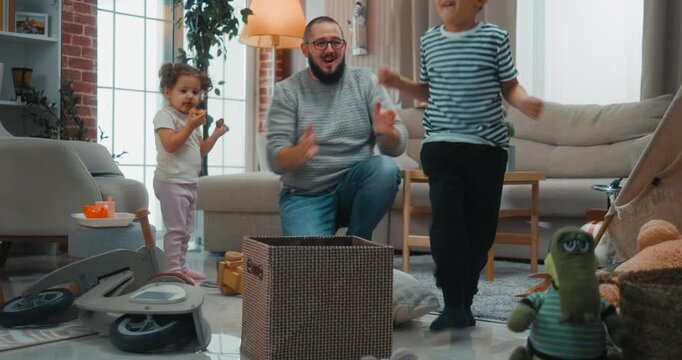 Single father motivates his kids to clean up the living room by turning toy pickup into a fun basketball game, throwing toys into a box. Positive parenting and playful everyday routine at home.