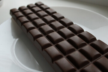 Close-up of dark or milk chocolate bar showing detailed rectangular segments and smooth surface texture on white plate background.