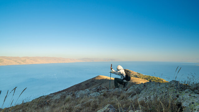 Hiker kneeling on a rocky mountain near Lake Sevan, Armenia, with trekking poles, looking at the calm blue lake and golden hills under clear sunlight.
