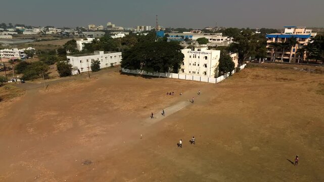 Aerial view of children playing cricket on a dry field, with buildings in the background, contrasting the open space, Patas, Maharashtra, India.