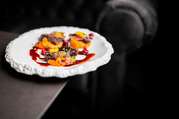 A finely plated gourmet dish featuring meat, a rich orange purée, and a dark sauce on a white textured plate in a dim, restaurant setting