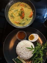 A bowl of shrimp in spicy coconut milk (udang lemak cili api) served alongside rice, sambal, boiled egg, cucumber, and ulam on a dark plate, showcasing vibrant Malaysian flavors. Top view.