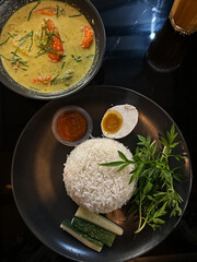 A bowl of shrimp in spicy coconut milk (udang lemak cili api) served alongside rice, sambal, boiled egg, cucumber, and ulam on a dark plate, showcasing vibrant Malaysian flavors. Top view.
