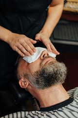 Fototapeta premium Bearded man relaxes in a barber chair while a hot towel is gently applied to his face as part of a grooming or skincare treatment
