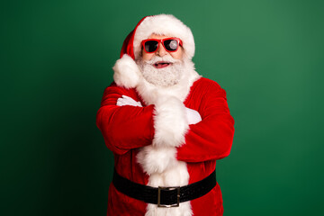 Santa Claus in red suit with white fur hat crosses arms wearing red sunglass look for a festive holiday vibe against a green backdrop