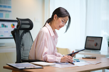 Young professional woman reviewing data on tablet and paper documents in a bright office.
