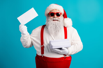 Santa in red suit with white beard wears hat suspenders and red glasses while holding a stack of...