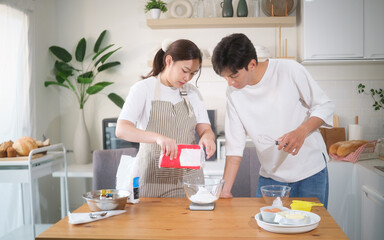 Couple preparing to bake together in a bright kitchen, woman measuring flour while man watches with whisk in hand. Concept of teamwork and home cooking.