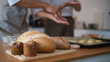 Close-up of freshly baked bread and muffins on a wooden board with blurred couple baking in the background
