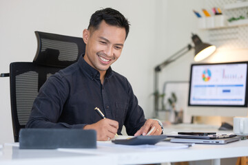 Smiling businessman working at desk in modern office, writing notes and analyzing financial reports on computer screen.