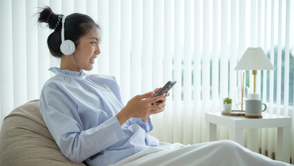 Asian woman sitting on a beanbag, wearing headphones and smiling while using her smartphone in a bright, modern room.
