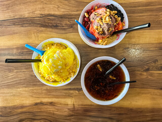 Three bowls of shaved ice desserts with vibrant toppings including chocolate ice cream, jelly, sweet corn, and syrup, served on a wooden café table.