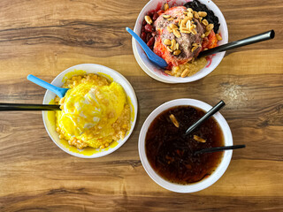 Three bowls of shaved ice desserts with vibrant toppings including chocolate ice cream, jelly, sweet corn, and syrup, served on a wooden café table.