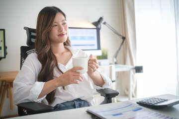 Asian woman enjoying a coffee break while working from home, seated at a bright and modern desk.