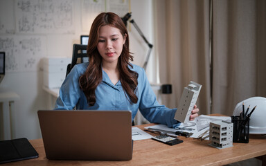 Female architect holding a building model while working on designs at her desk.