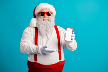 Santa in red suspenders with beard and funny look on bright blue background holds a phone promoting...