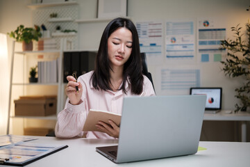 Woman focused on work, taking notes while analyzing data on her laptop in a modern office.