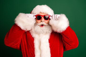 Santa in a red suit with white fur trim wears bright red sunglass against a green background for a festive playful holiday image