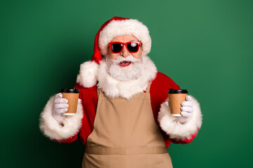 Santa in festive costume with hat beard and sunglass holding coffee cups in front of green background