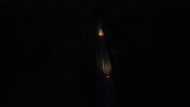 Aerial view of vehicles traveling along winding road at night, illuminated headlights cutting through darkness, showcasing dynamic motion and depth, camera follows action with smooth tracking shot