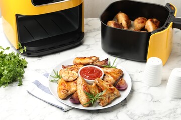 Delicious grilled chicken wings, sauce and spices near air fryer with cooked drumsticks on white marble table