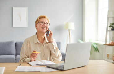 Smiling adult woman with short blonde hair wearing glasses sitting at desk with laptop and talking...