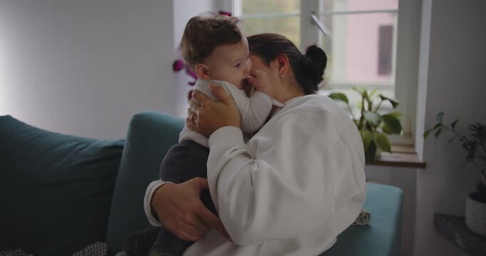 Mother cuddling baby affectionately in living room by window, child resting against her as they share tender moment of calm and connection