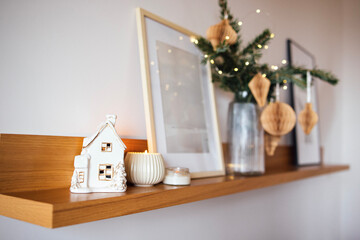 Cozy home decor featuring a white ceramic house, candles, and framed artwork on a wooden shelf, adorned with festive greenery and decorative ornaments for a warm atmosphere