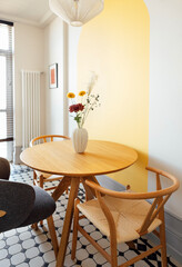A stylish dining room with a round wooden table with a vase and comfortable chairs made of natural materials. Contrasting interior with white walls and bright yellow details.