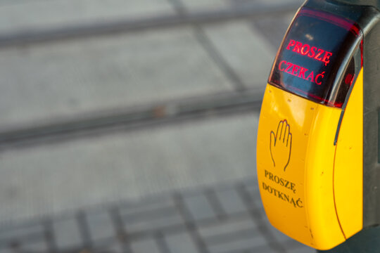 Pedestrian crossing button. Yellow device displaying PROSZĘ CZEKAĆ and PROSZĘ DOTKNĄĆ. Detail of urban traffic light infrastructure and street environment.