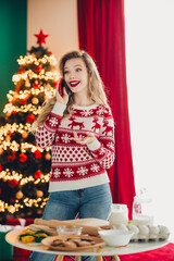 Cheerful woman in a cozy holiday sweater chats on the phone by a twinkling christmas tree with...