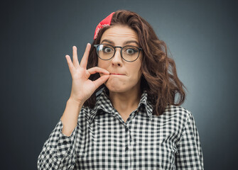 Keep the secret - keep quiet. Young woman keeps closed mouth with her fingers over dark gray -...