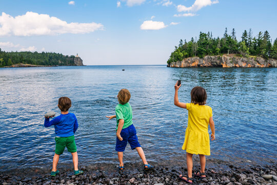Rear view of Three young children standing side by side at the edge of a lake throwing rocks into the water in summer