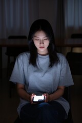 In the darkness, a girl looks at the screen of a mobile phone and sadly waits for a call, her face illuminated by the light from the phone.