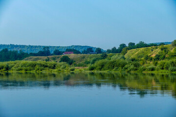 picturesque riverbank, green slope, reflection in water, roofs of houses, hills, mountains in distance, forest, summer greenery, meadow grass, hike, travel, tourism, leisure, horizontal photo