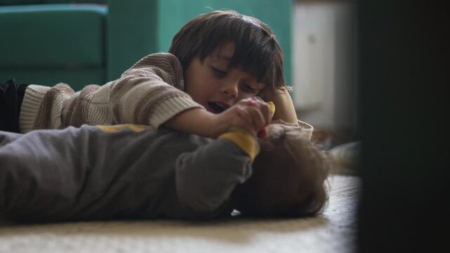 Older brother lying on floor and playing gently with baby sibling, both smiling and holding hands, expressing joy, affection, and warmth in tender family moment at home