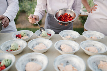 Chef prepares dessert outdoors, adding fresh strawberries to bowls of green and pink ice cream. Several servings ready on the table, creating a fresh, summery vibe. Ideal for food styling or event pro