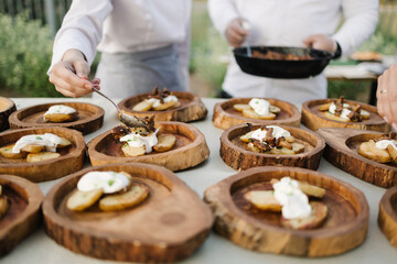 Chefs prepare rustic appetizers on wooden serving boards outdoors. Sliced potatoes topped with creamy sauce and garnished with herbs. One chef adds final touches with a spoon, another holds a pan with