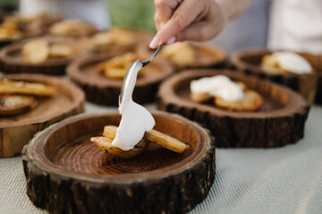 A hand adds creamy sauce to roasted potato slices on a rustic wooden plate. Close-up of outdoor catering setup with multiple similar plates in the background, perfect for event food imagery.