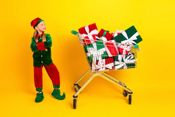 Festive young elf dressed in vibrant costume posing near cart of gift boxes in cheerful yellow ambiance celebrating holidays