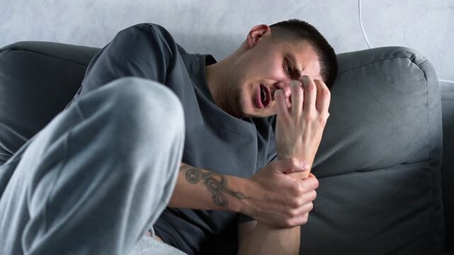 A young man with a tattoo on his arm massages his wrist while sitting on a gray sofa.
Arm pain due to injury.
Forearm bruise.
Pain, arm, bruise, forearm, injury, massage.