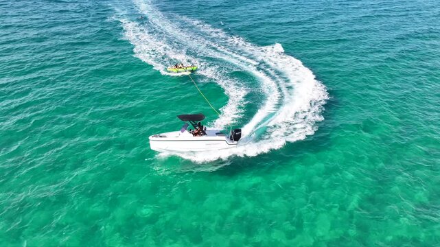 Aerial drone tracking video of powerboat towing inflatable tubes used for water sports in tropical exotic bay with emerald crystal clear sea