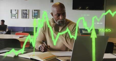 Senior exec in tan sweater writing with pen reviewing laptop at coworking space, with chart overlay