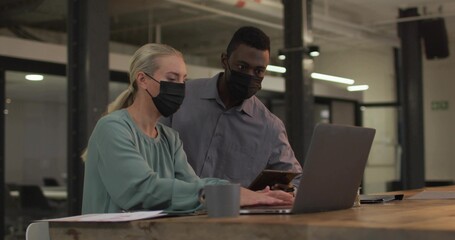 Collaborating masked colleagues examining smartphone typing at office desk with laptop coffee mug