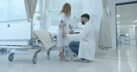 Kneeling doctor helping child in gown using crutches in ward, with bed, IV pole, cart, curtains
