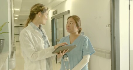 Pointing doctor in lab coat guiding nurse viewing digital tablet in hospital hall, with stethoscope